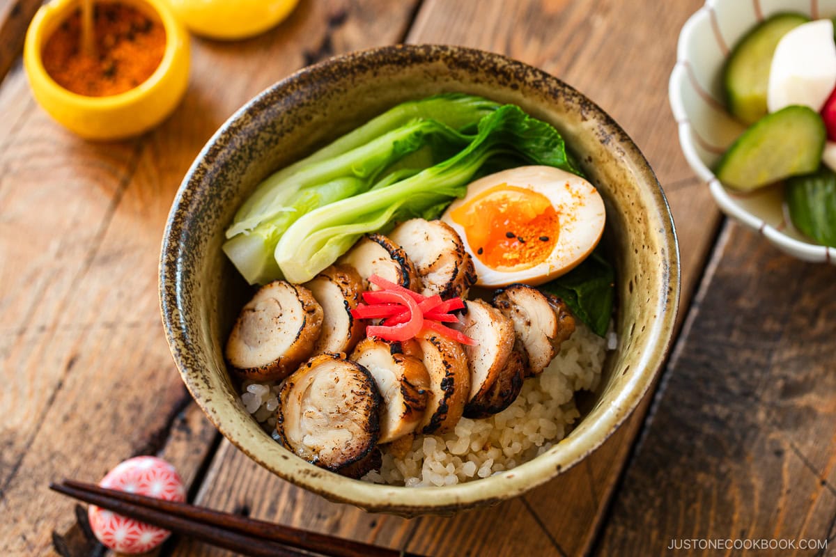 A donburi bowl containing steamed rice topped with seared sliced chicken chashu, soft boiled egg, and blanched bok choy, garnished with pickled ginger.
