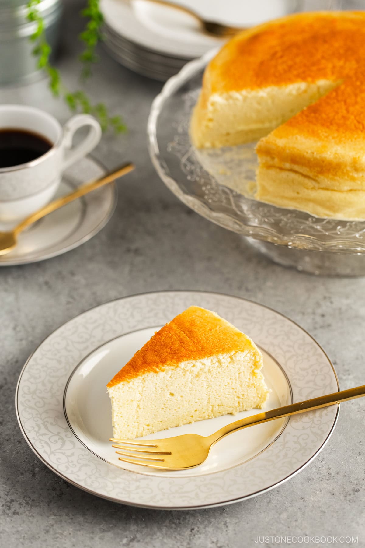 A white and gray rim plate containing a slice of Japanese souffle cheesecake. A cake stand behind containing the whole cake.