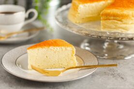 A white and gray rim plate containing a slice of Japanese souffle cheesecake. A cake stand behind containing the whole cake.
