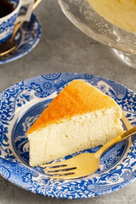 A blue and white plate containing a slice of Japanese souffle cheesecake. A cake stand behind containing the whole cake.