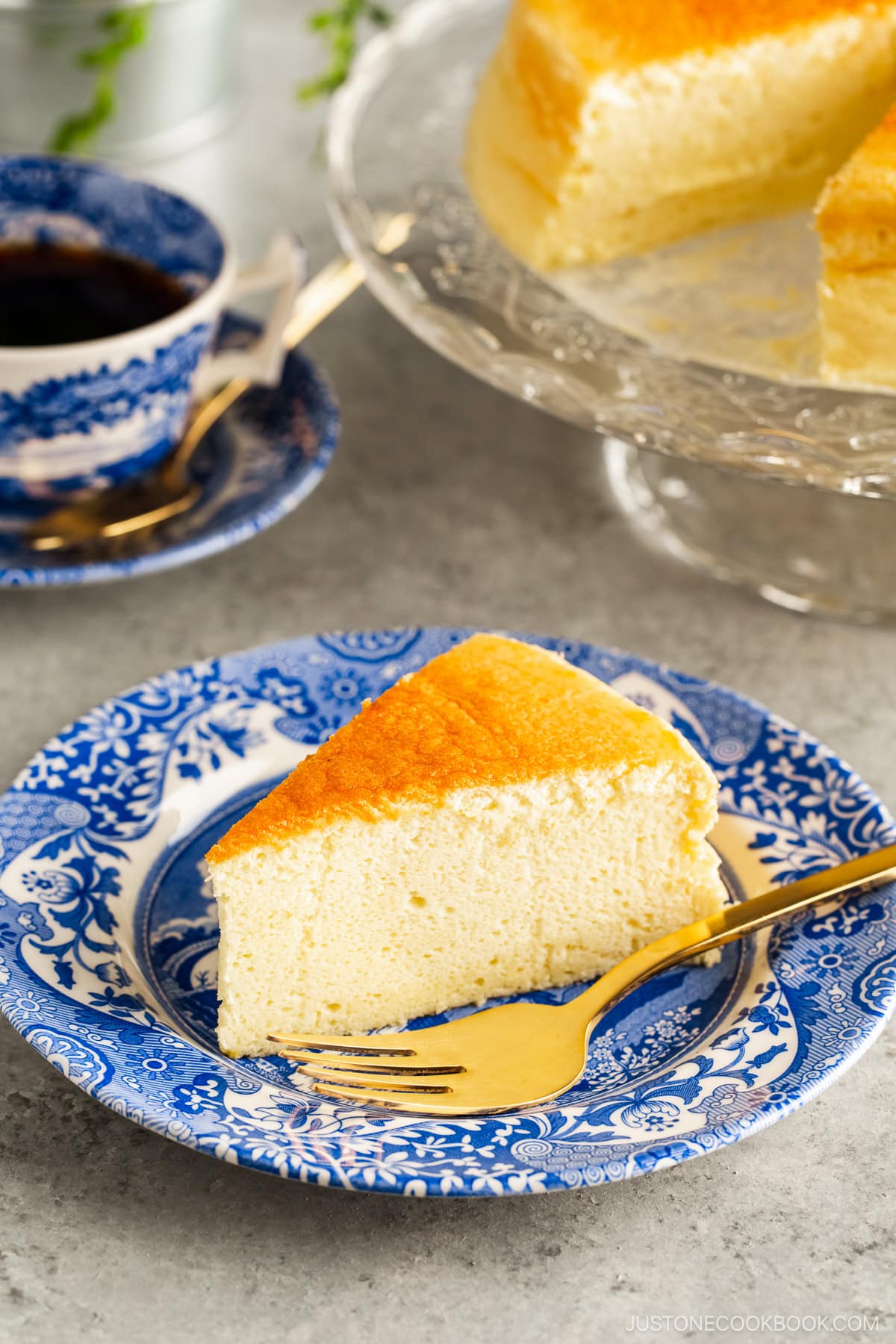 A blue and white plate containing a slice of Japanese souffle cheesecake. A cake stand behind containing the whole cake.