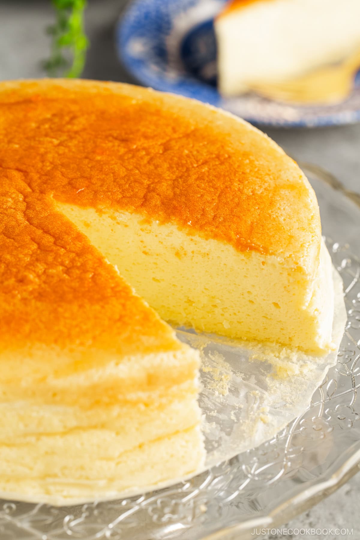 A glass cake stand containing Japanese souffle cheesecake slice being cut out and served onto a blue and white plate.