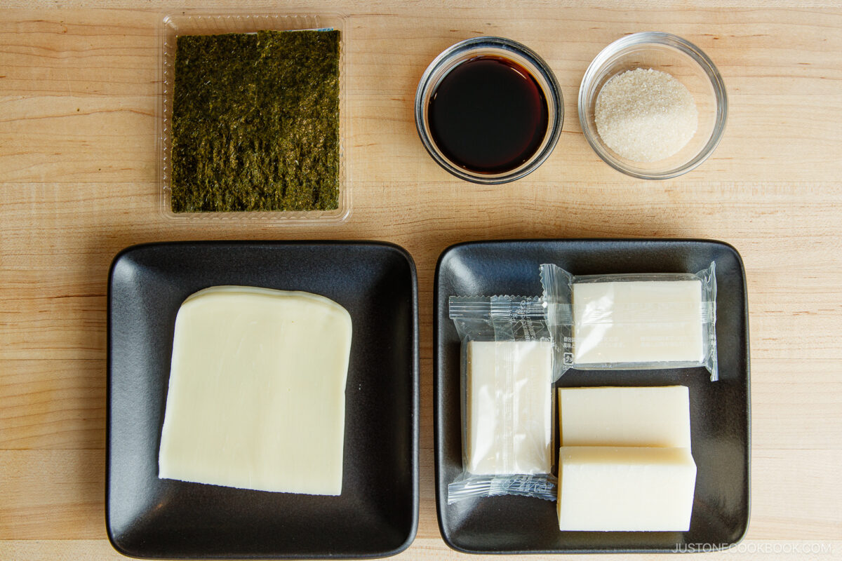 Nori seaweed sheet, soy sauce, sugar, slices of tofu, and packaged tofu blocks arranged on black plates, ideal for preparing authentic Japanese tofu dish or appetizer, with traditional ingredients for Japanese cuisine.