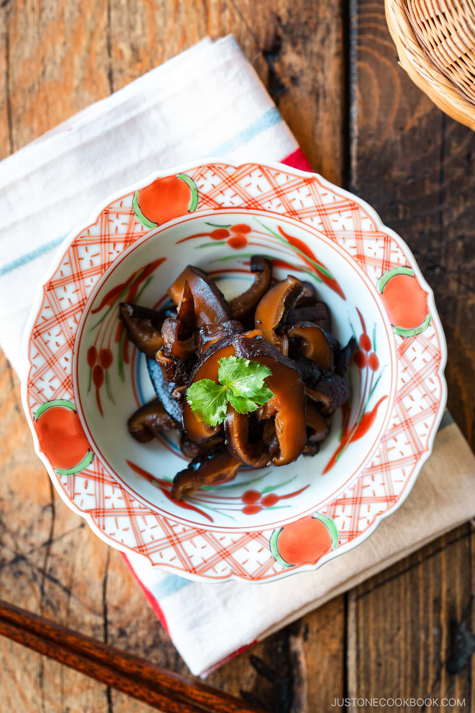 A round red bowl containing Simmered Shiitake Mushrooms (Fukumeni) garnished with green leaves.