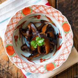 A round red bowl containing Simmered Shiitake Mushrooms (Fukumeni) garnished with green leaves.