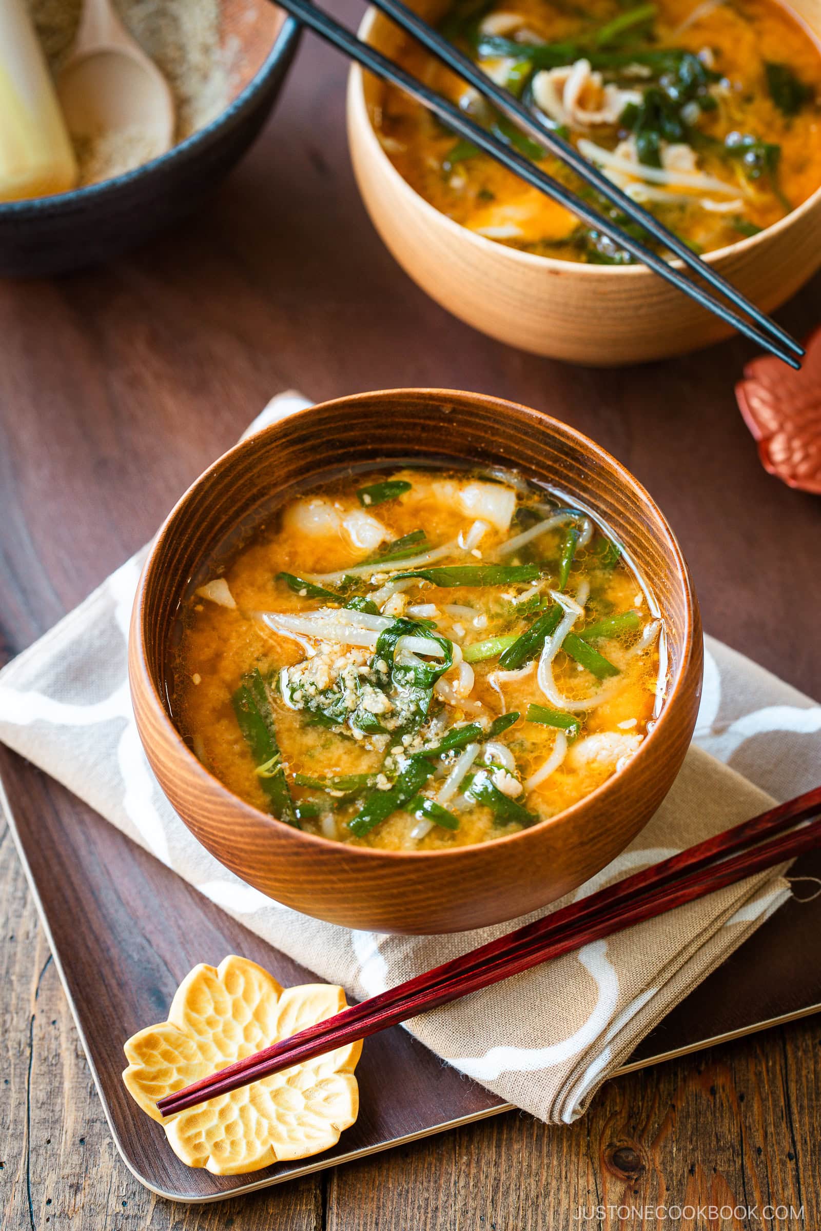 Light and dark-colored wooden bowls containing Spicy Pork and Bean Sprout Miso Soup sprinkled with ground sesame seeds, placed on a wooden the table.
