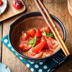 A Japanese black mortar containing cut tomato wedges dressed in ground sesame ponzu seasoning, garnished with mitsuba leaves on top.