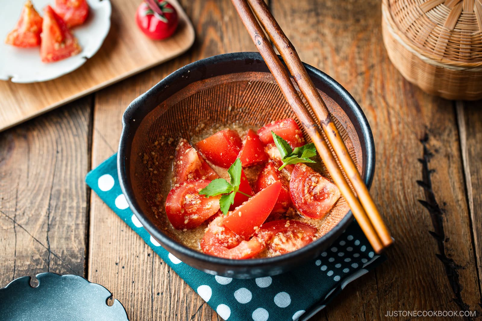A Japanese black mortar containing cut tomato wedges dressed in ground sesame ponzu seasoning, garnished with mitsuba leaves on top.
