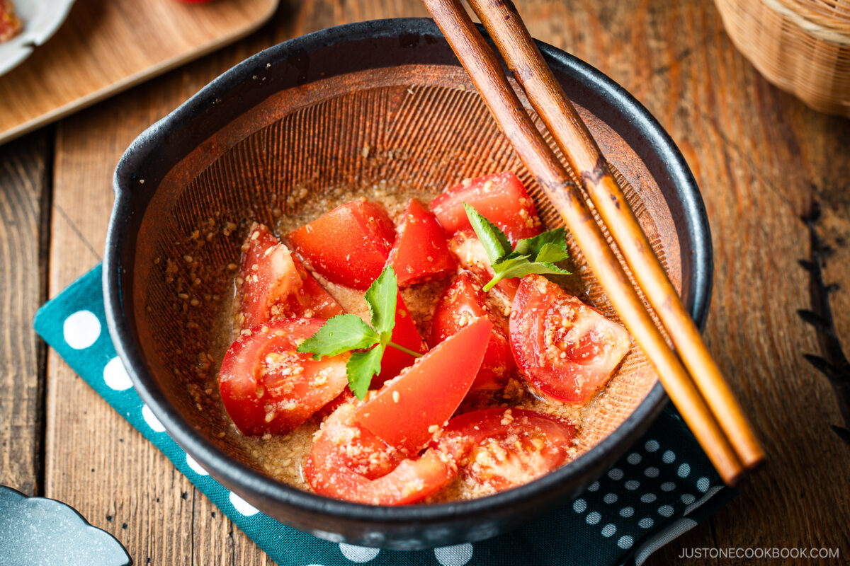 A Japanese black mortar containing cut tomato wedges dressed in ground sesame ponzu seasoning, garnished with mitsuba leaves on top.