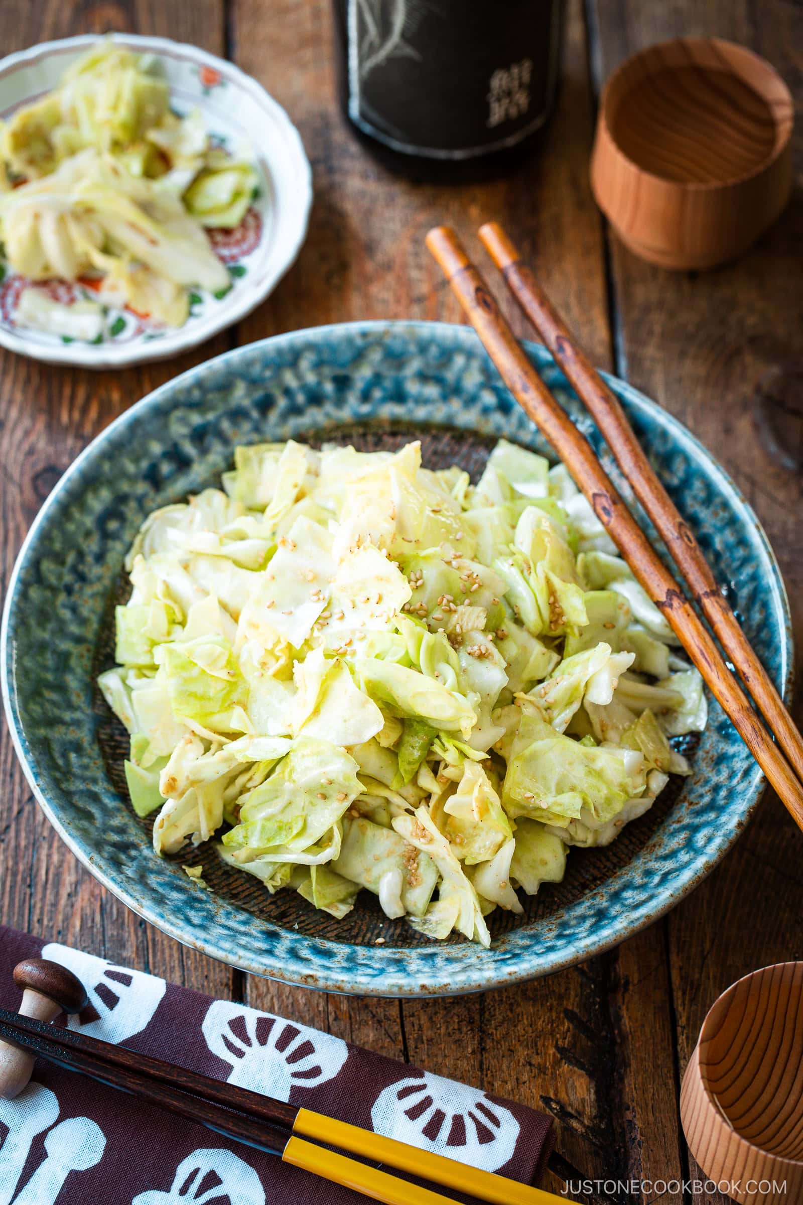 An indigo round plate containing chopped cabbage dish called Yamitsuki Cabbage served with a pair of chopsticks and small dishes and sake cups and the table.