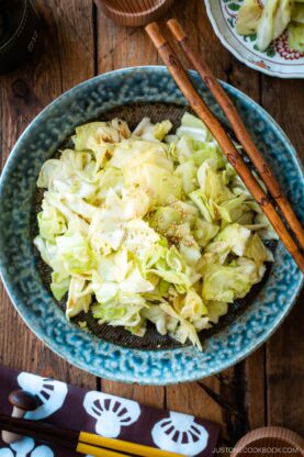 An indigo round plate containing chopped cabbage dish called Yamitsuki Cabbage served with a pair of chopsticks and small dishes and sake cups and the table.