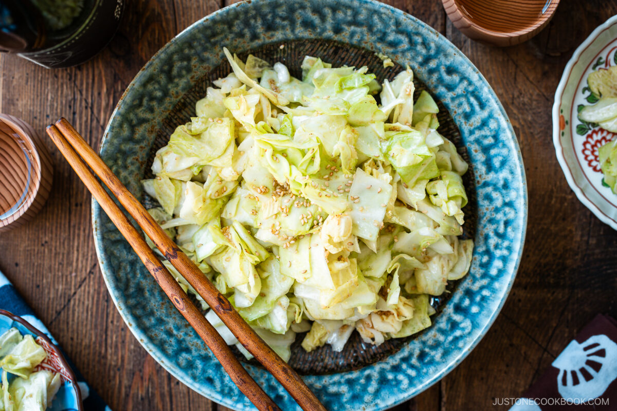 An indigo round plate containing chopped cabbage dish called Yamitsuki Cabbage served with a pair of chopsticks and small dishes and sake cups and the table.