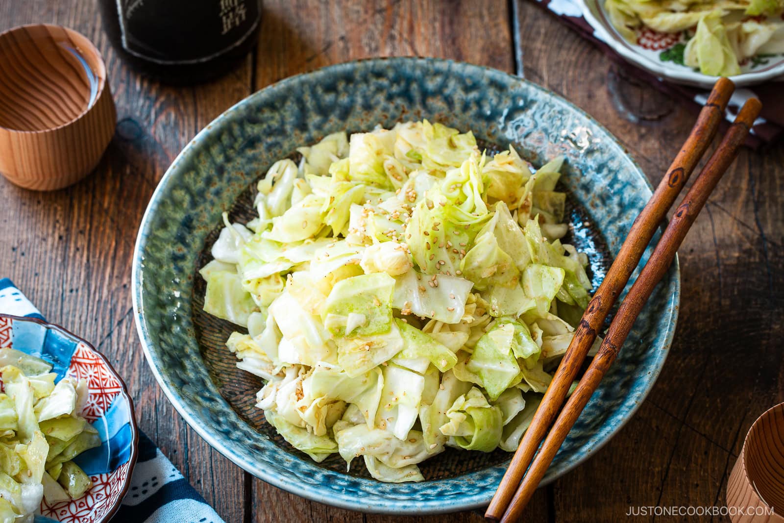 An indigo round plate containing chopped cabbage dish called Yamitsuki Cabbage served with a pair of chopsticks and small dishes and sake cups and the table.