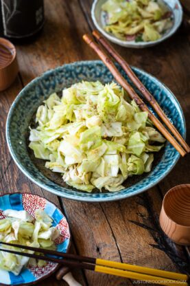 An indigo round plate containing chopped cabbage dish called Yamitsuki Cabbage served with a pair of chopsticks and small dishes and sake cups and the table.