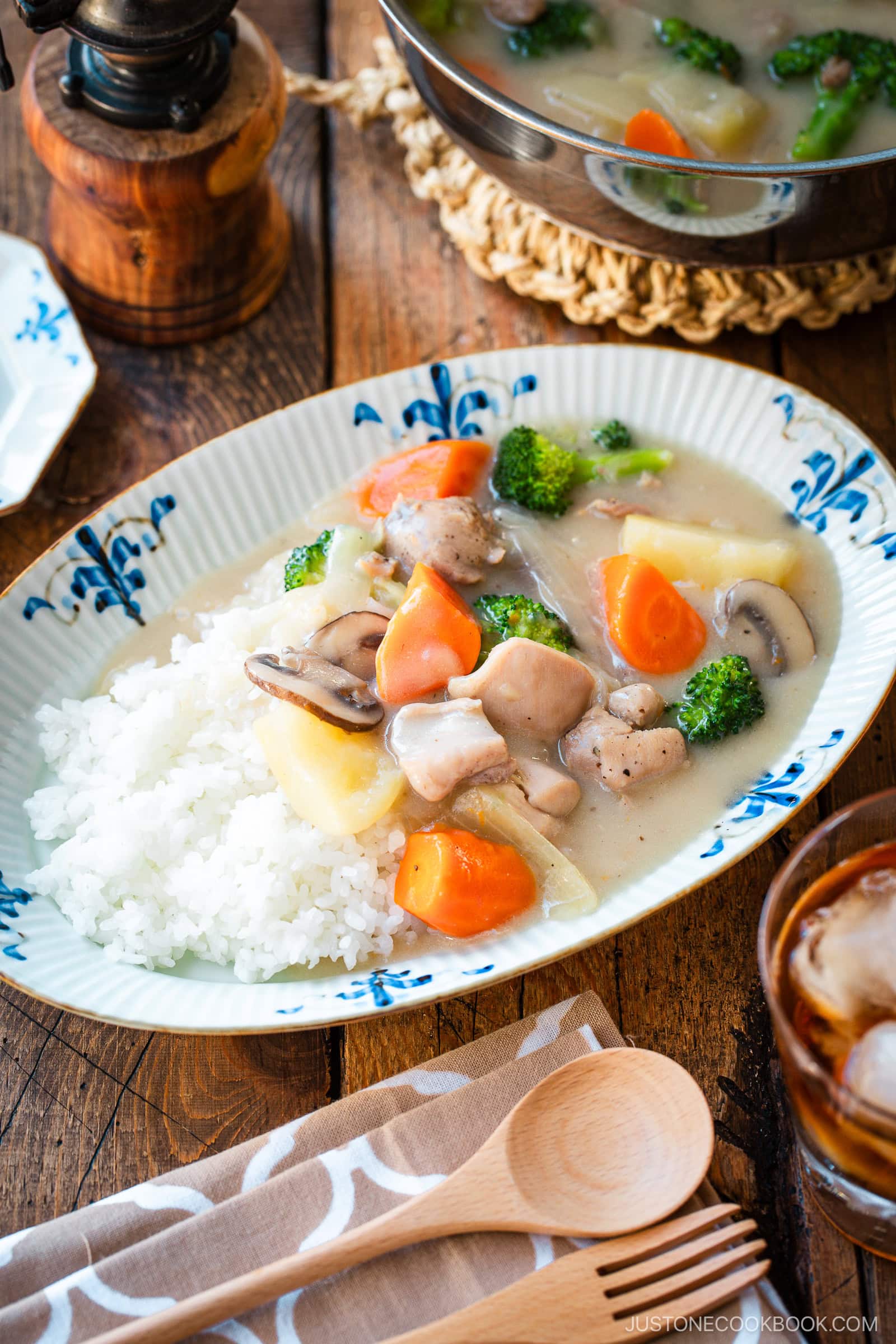 An oval-shaped plate holds a comforting serving of Japanese cream stew, featuring tender chicken, carrots, potatoes, mushrooms, and broccoli in a rich, creamy white sauce. The stew is generously ladled over a bed of fluffy steamed rice, creating a hearty and satisfying meal.
