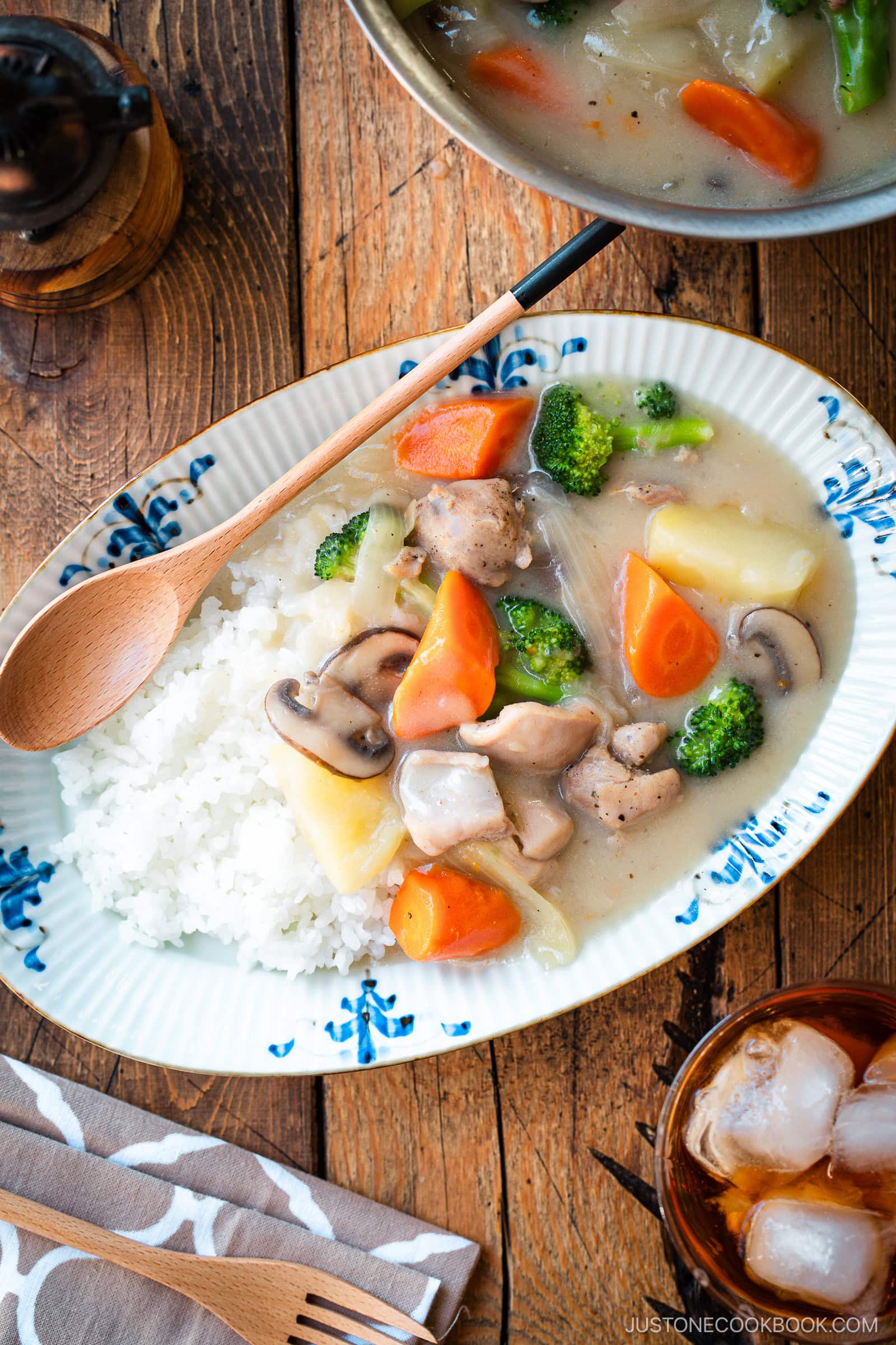 An oval-shaped plate holds a comforting serving of Japanese cream stew, featuring tender chicken, carrots, potatoes, mushrooms, and broccoli in a rich, creamy white sauce. The stew is generously ladled over a bed of fluffy steamed rice, creating a hearty and satisfying meal.