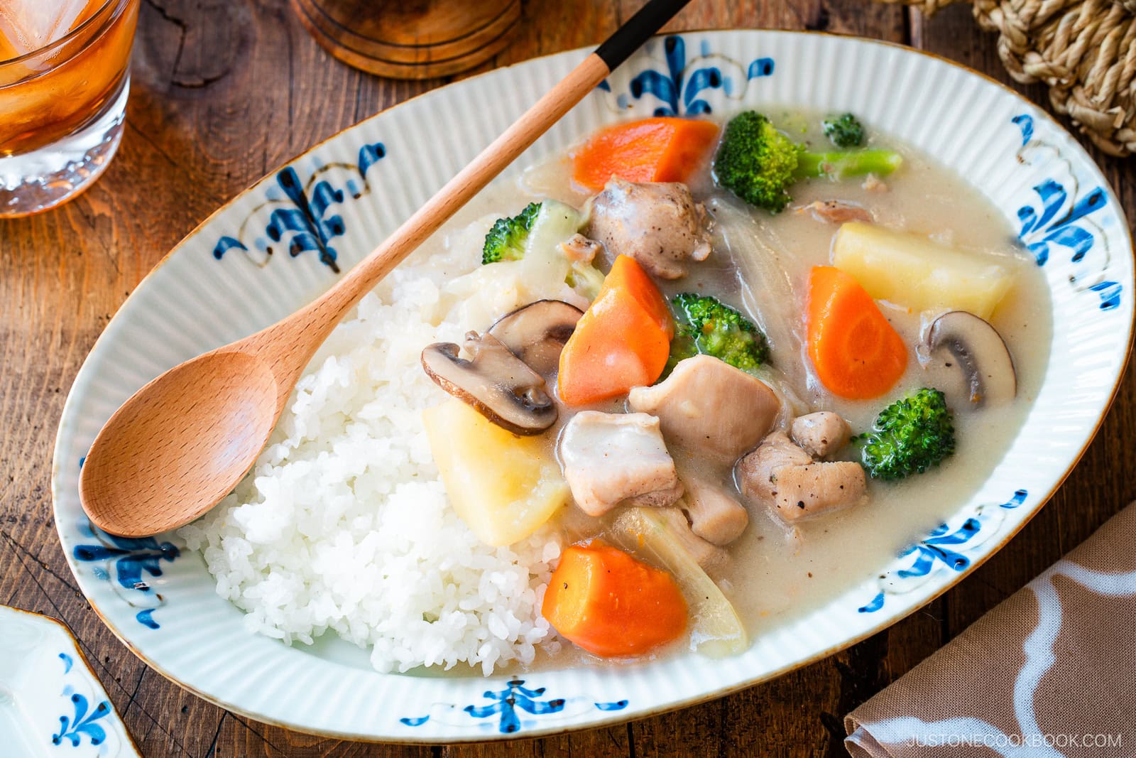 An oval-shaped plate holds a comforting serving of Japanese cream stew, featuring tender chicken, carrots, potatoes, mushrooms, and broccoli in a rich, creamy white sauce. The stew is generously ladled over a bed of fluffy steamed rice, creating a hearty and satisfying meal.