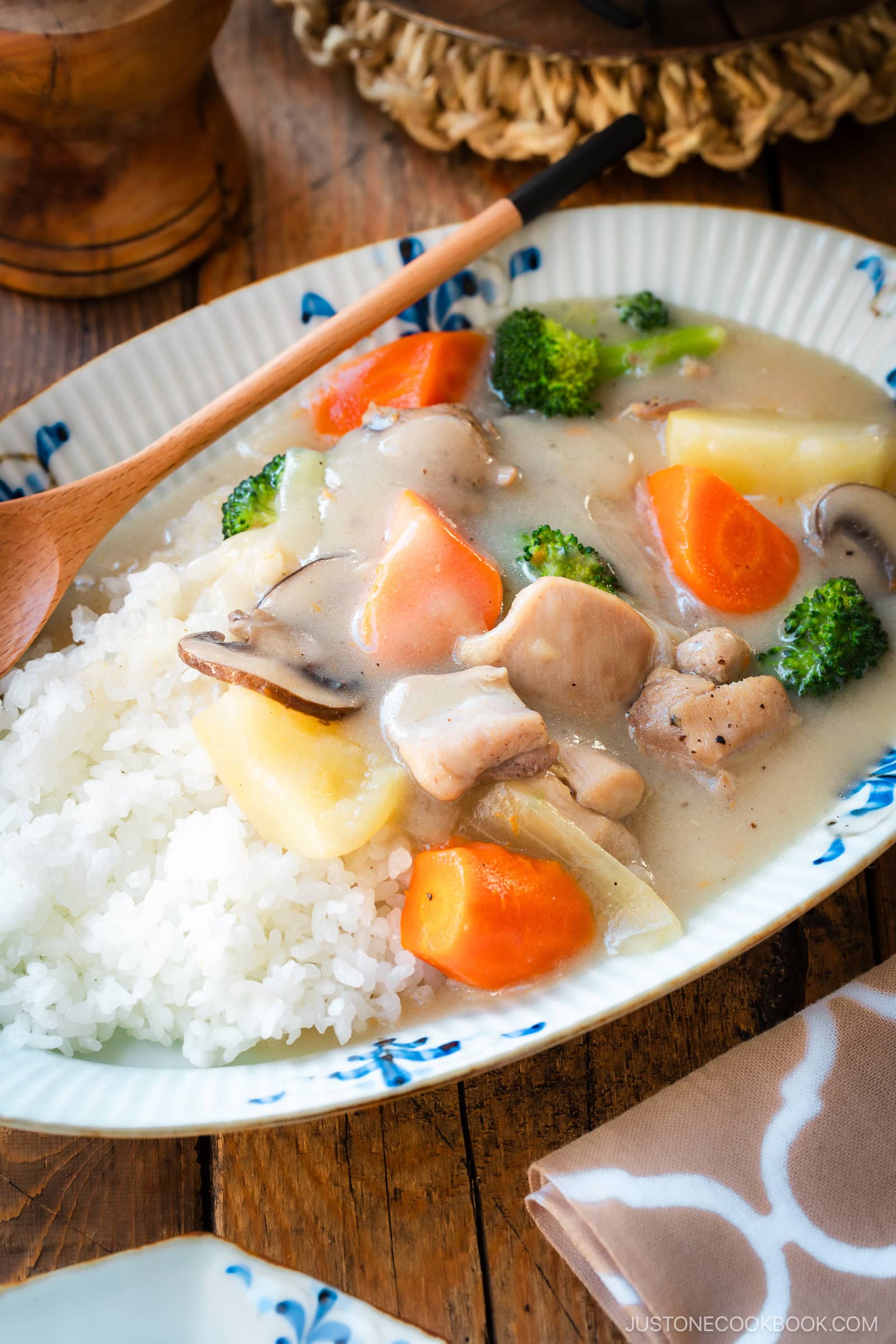 An oval-shaped plate holds a comforting serving of Japanese cream stew, featuring tender chicken, carrots, potatoes, mushrooms, and broccoli in a rich, creamy white sauce. The stew is generously ladled over a bed of fluffy steamed rice, creating a hearty and satisfying meal.