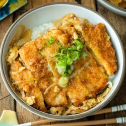 A white bowl containing Katsudon, pork cutlet rice bowl, where golden brown tonkatsu, drizzled egg, and tender onion are simmered in a savory dashi broth.