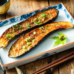 A rectangular plate containing Miso Glazed Eggplant garnished with yuzu kosho paste on a green maple leaf on the side.