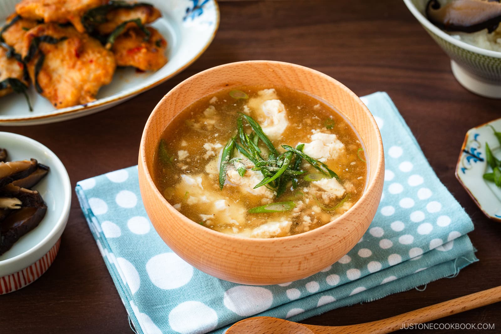 Wooden bowls filled with Pork Tofu Soup, served alongside steamed rice and a chicken dish on the table.