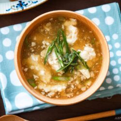 Wooden bowls filled with Pork Tofu Soup, served alongside steamed rice and a chicken dish on the table.