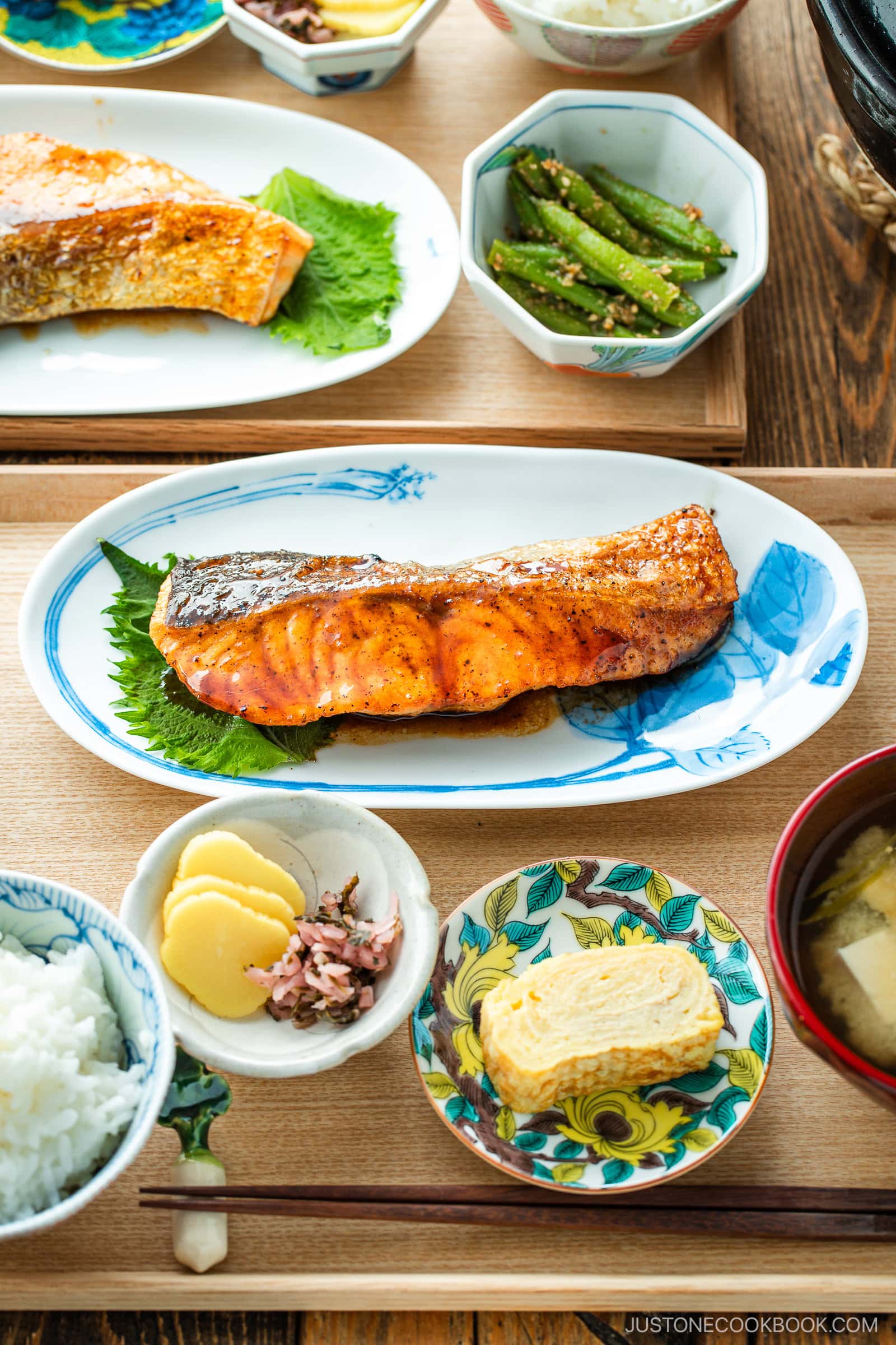 A Japanese meal consisting of small dishes or bowls, each plating Teriyaki Salmon, steamed rice, miso soup, pickles, tamagoyaki, and green bean gomaae.