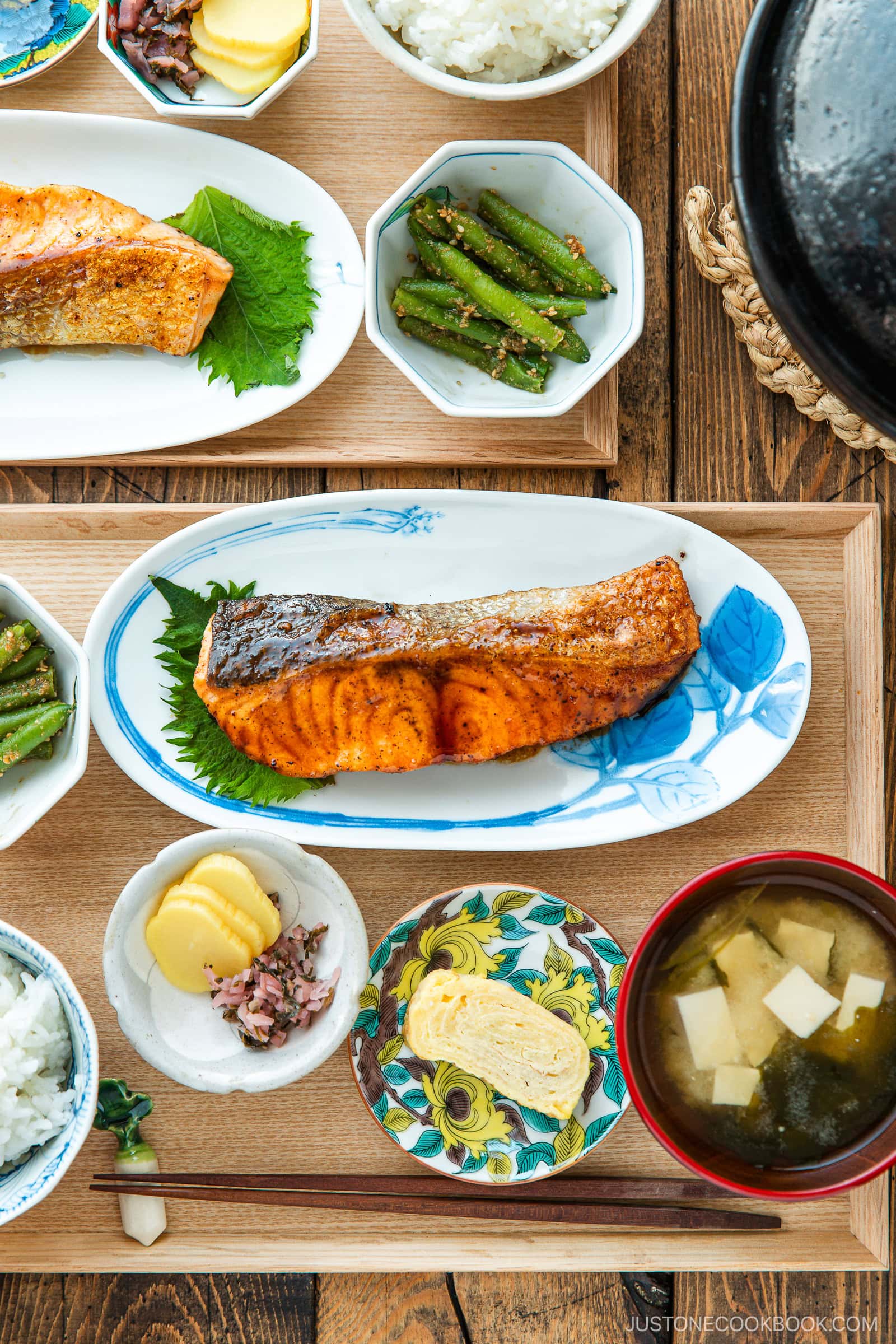 A Japanese meal consisting of small dishes or bowls, each plating Teriyaki Salmon, steamed rice, miso soup, pickles, tamagoyaki, and green bean gomaae.