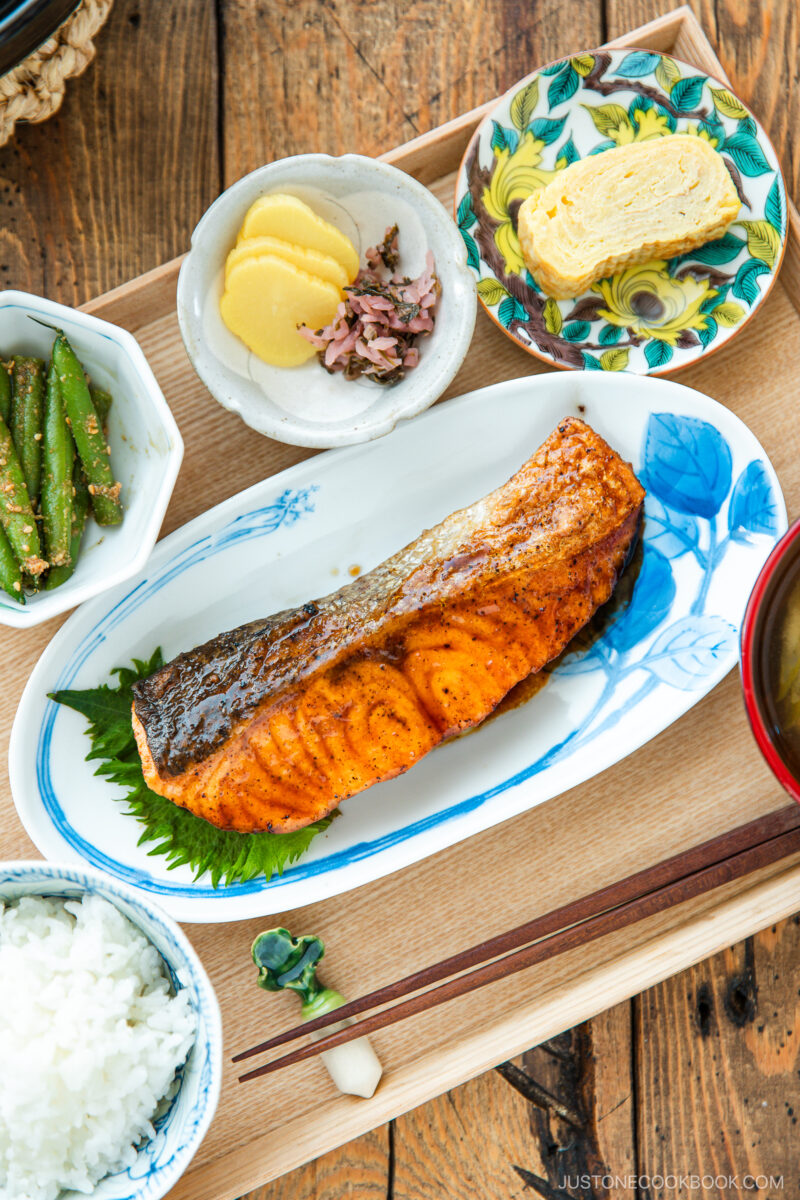 A Japanese meal consisting of small dishes or bowls, each plating Teriyaki Salmon, steamed rice, miso soup, pickles, tamagoyaki, and green bean gomaae.