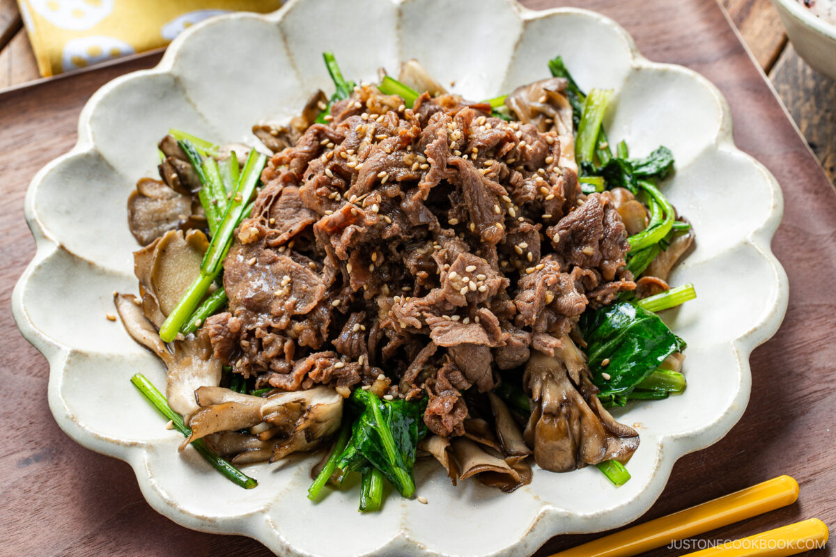 A white scalloped plate containing stir-fried Butter Ponzu beef with komatsuna green and maitake mushrooms.