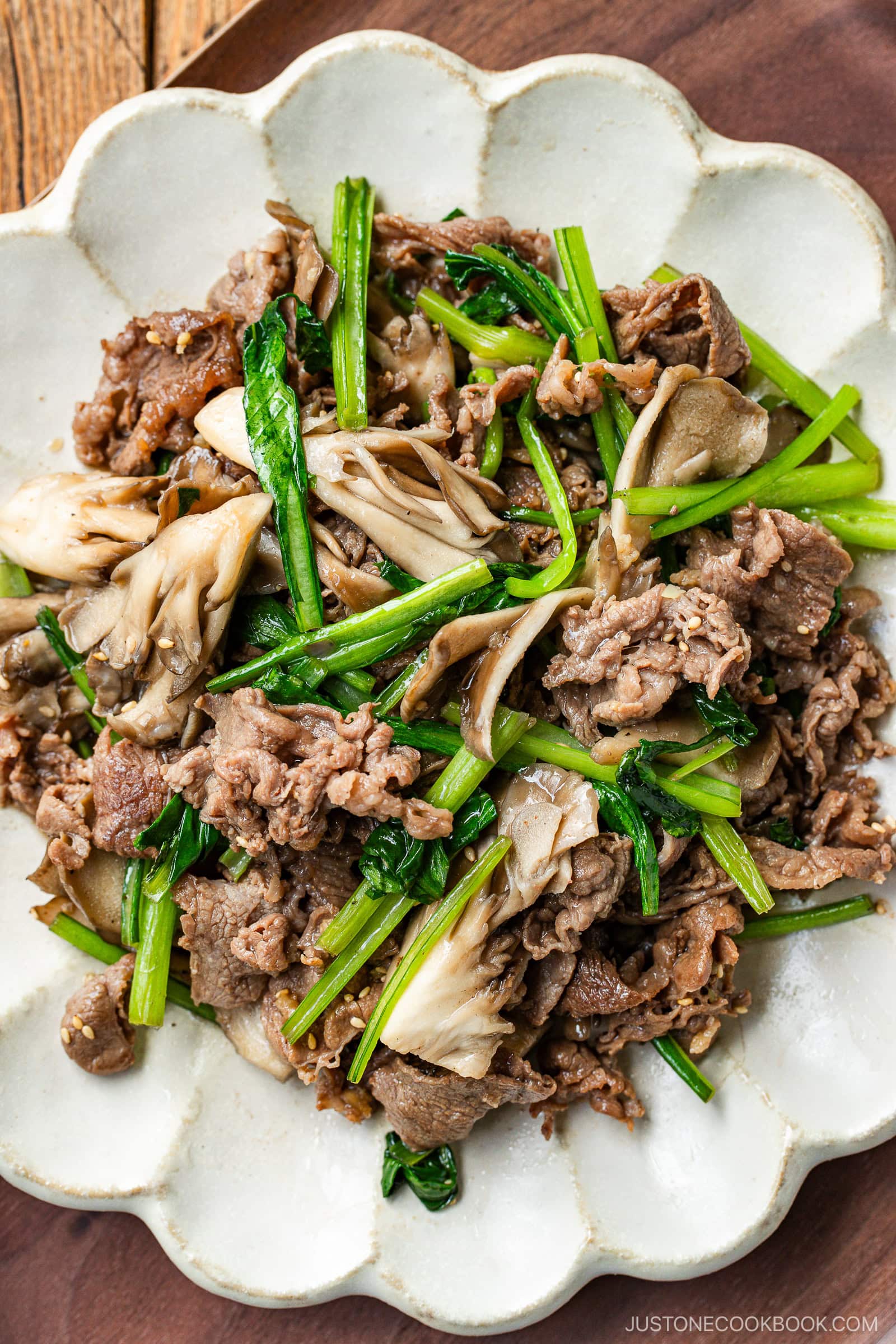 A white scalloped plate containing stir-fried Butter Ponzu beef with komatsuna green and maitake mushrooms.