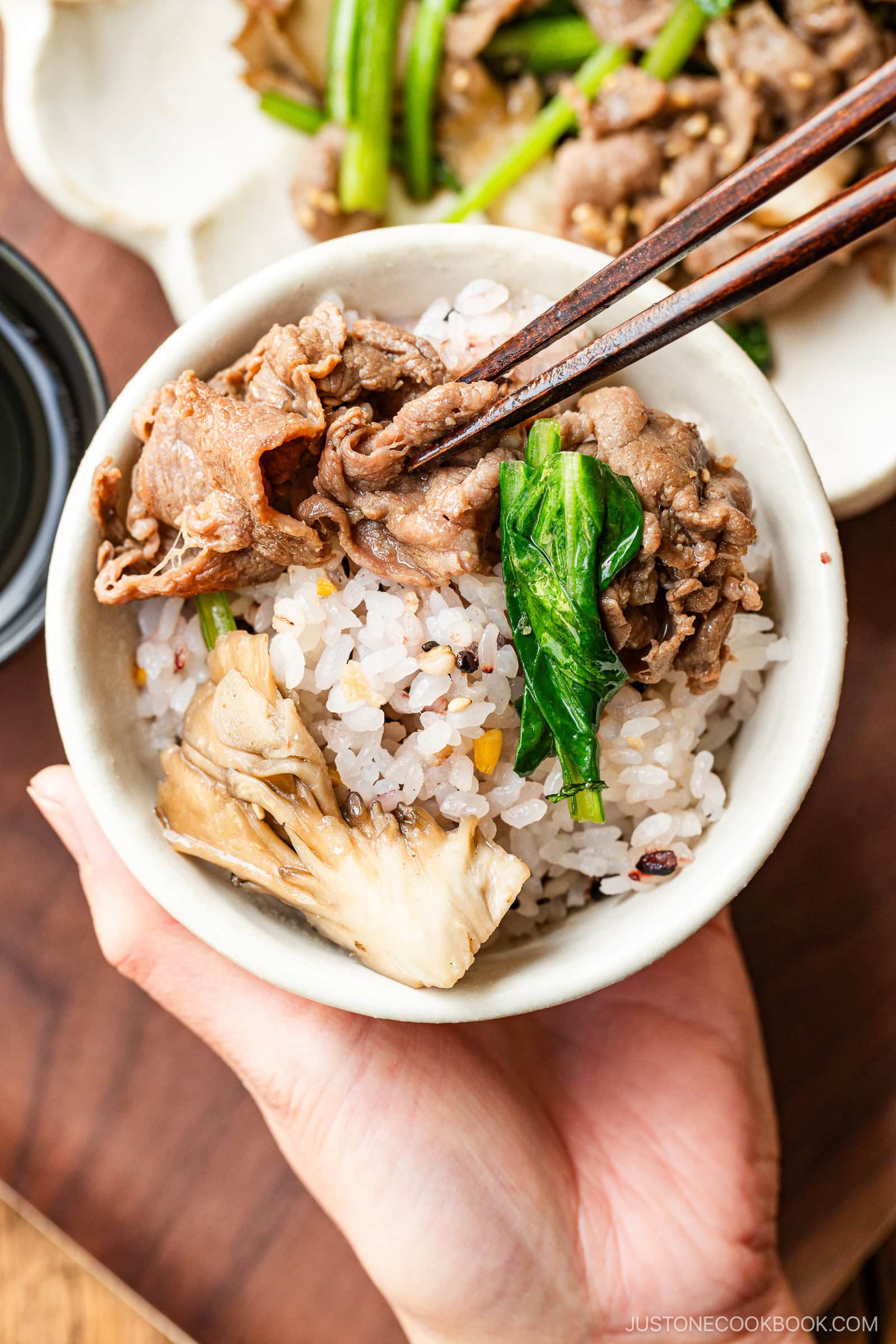 A white rice bowl containing multigrain rice and Butter Ponzu Beef with leafy green and Japanese wild mushrooms.