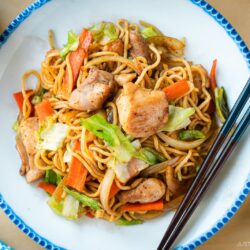 Delicious Chicken Yakisoba with tender, moist chicken thighs and crisp veggies in a sweet savory sauce plated on a beautiful white plate with blue scalloped edge.