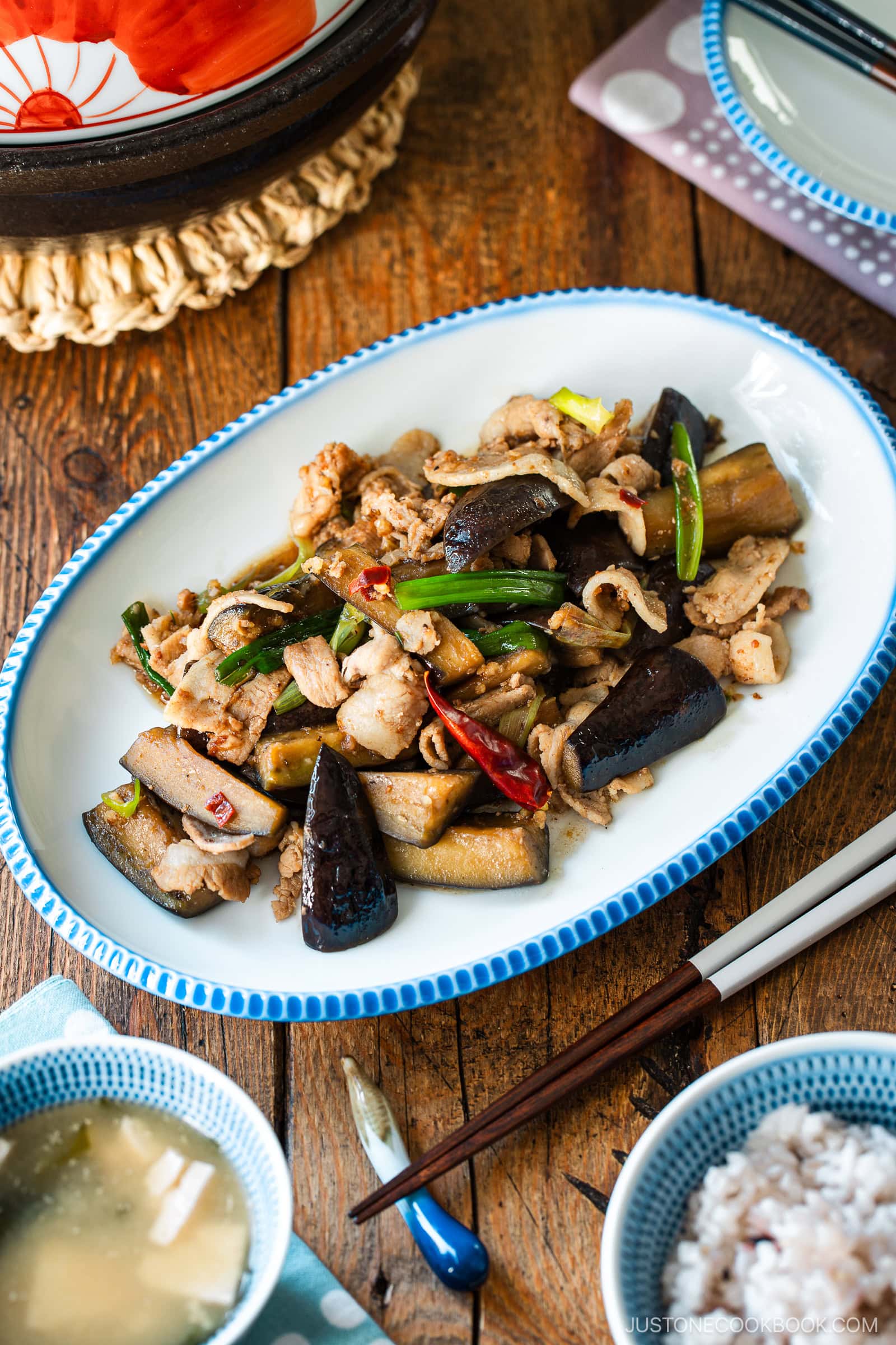 An oval plate with scalloped edge containing Pork and Eggplant Stir Fry served with multigrain rice and miso soup.