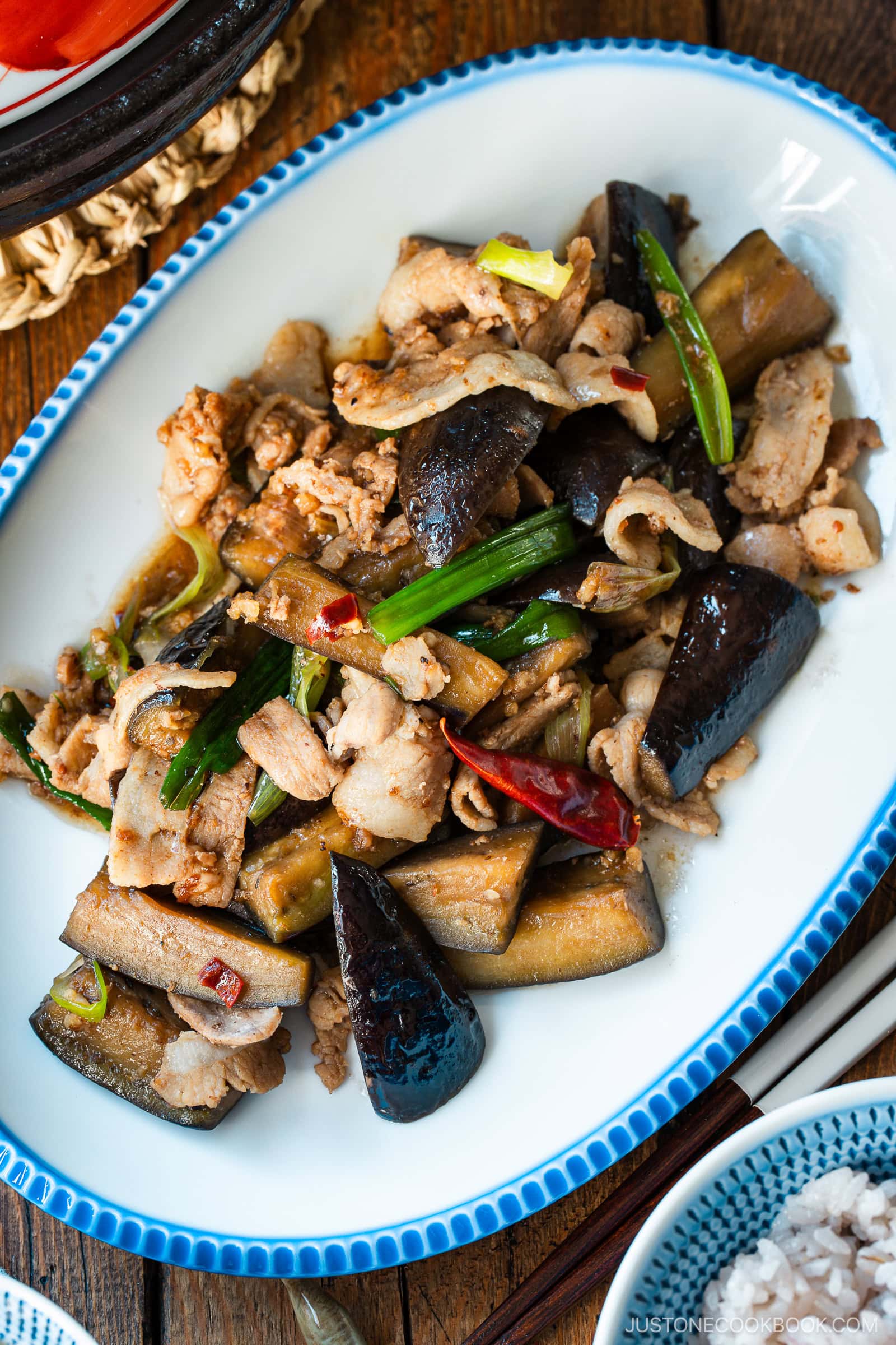 An oval plate with scalloped edge containing Pork and Eggplant Stir Fry served with multigrain rice.