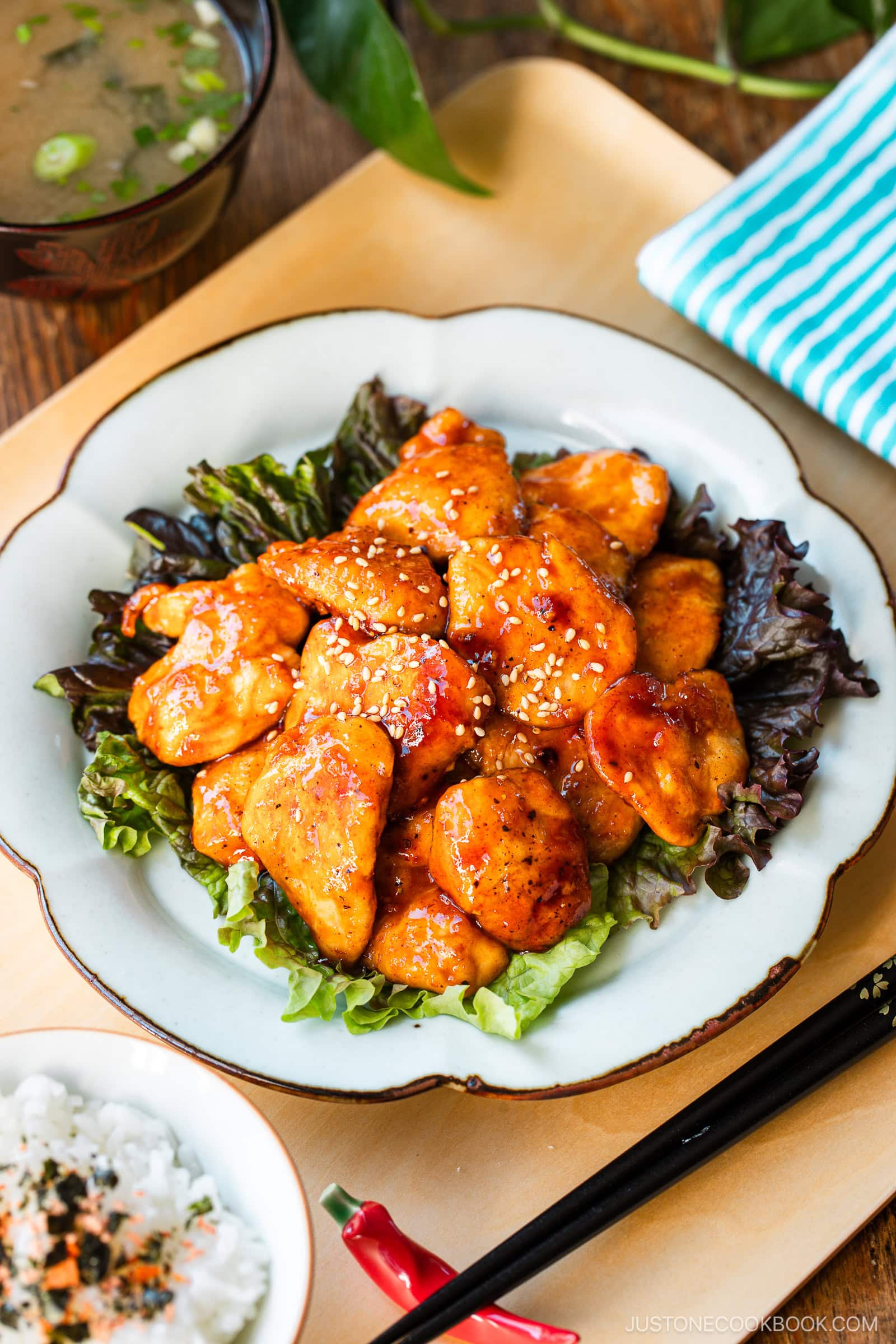 A white plate containing Honey Gochujang Chicken sprinkled with sesame chicken served on green salad.