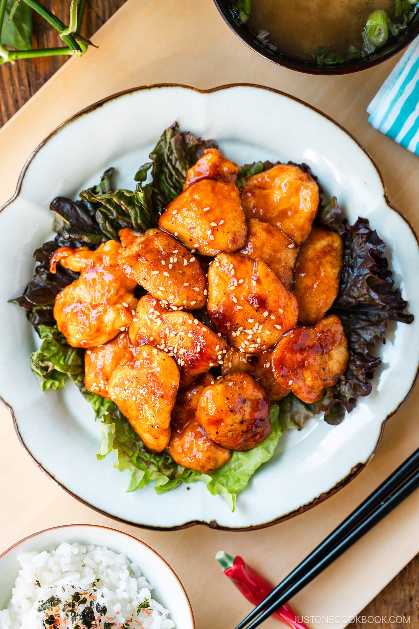 A white plate containing Honey Gochujang Chicken sprinkled with sesame chicken served on green salad.