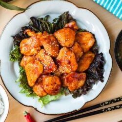 A white plate containing Honey Gochujang Chicken sprinkled with sesame chicken served on green salad.