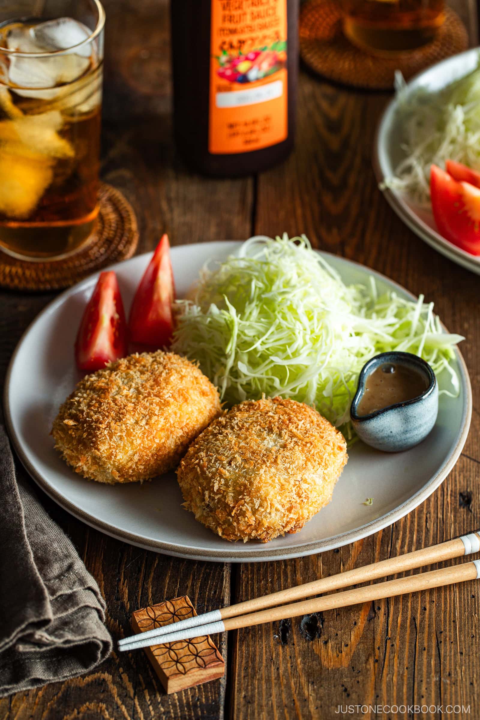 A round white plate containing Japanese croquettes, shredded cabbage, tomato wedges, and a tiny container with sesame dressing.