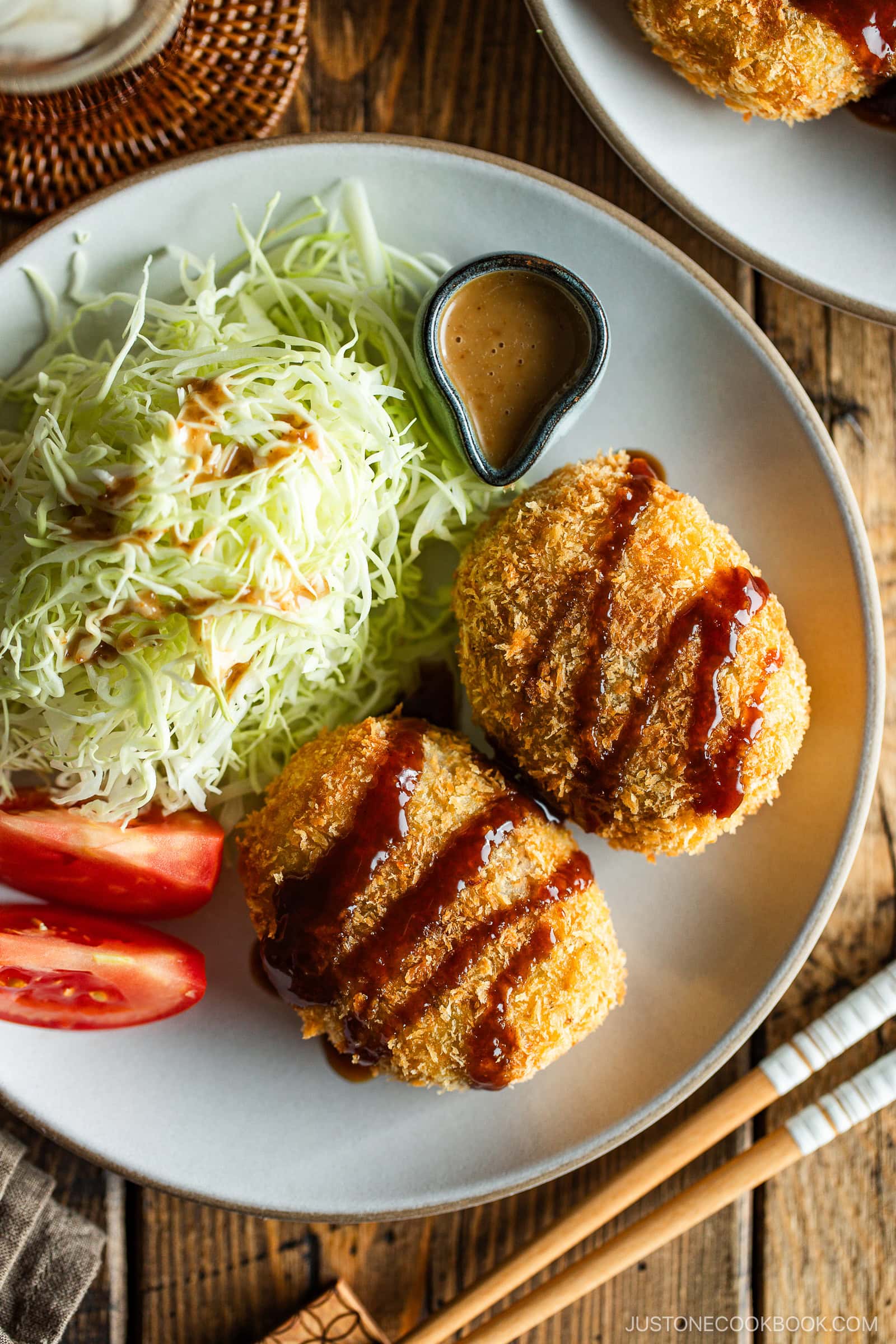 A round white plate containing Japanese croquettes, shredded cabbage, tomato wedges, and a tiny container with sesame dressing.