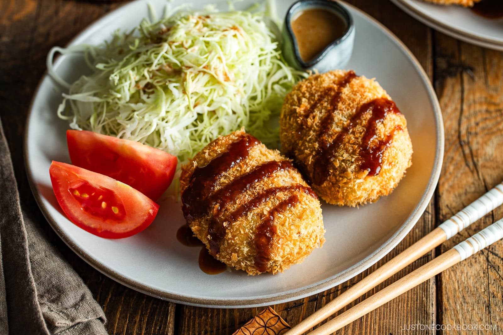 A round white plate containing Japanese croquettes, shredded cabbage, tomato wedges, and a tiny container with sesame dressing.