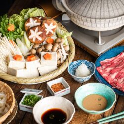 A Japanese shabu-shabu hot pot setup with a donabe pot, thinly sliced beef, tofu, assorted mushrooms, vegetables, udon noodles, and dipping sauces arranged on a wooden table.