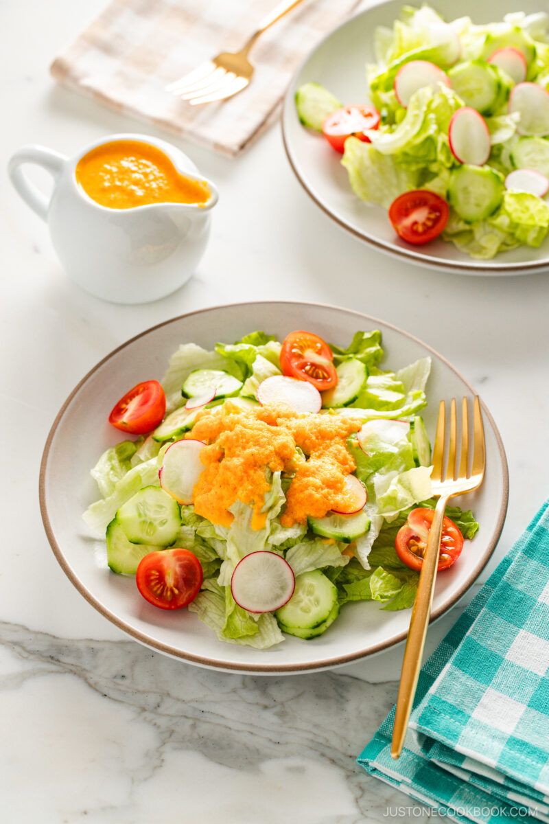 White plates of fresh green salad with lettuce, cucumbers, radishes, and cherry tomatoes, topped with vibrant orange carrot ginger dressing and served with a gold fork on a marble surface.