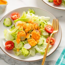 White plates of fresh green salad with lettuce, cucumbers, radishes, and cherry tomatoes, topped with vibrant orange carrot ginger dressing and served with a gold fork on a marble surface.
