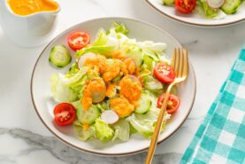 White plates of fresh green salad with lettuce, cucumbers, radishes, and cherry tomatoes, topped with vibrant orange carrot ginger dressing and served with a gold fork on a marble surface.