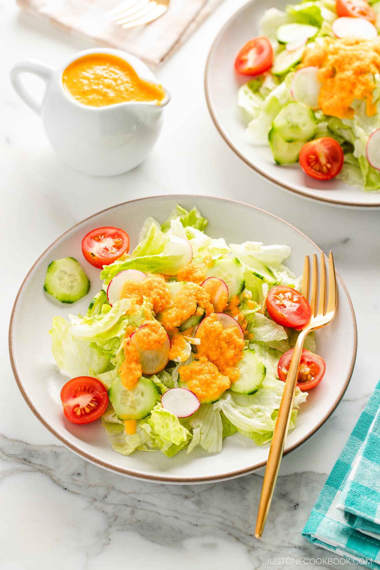 White plates of fresh green salad with lettuce, cucumbers, radishes, and cherry tomatoes, topped with vibrant orange carrot ginger dressing and served with a gold fork on a marble surface.