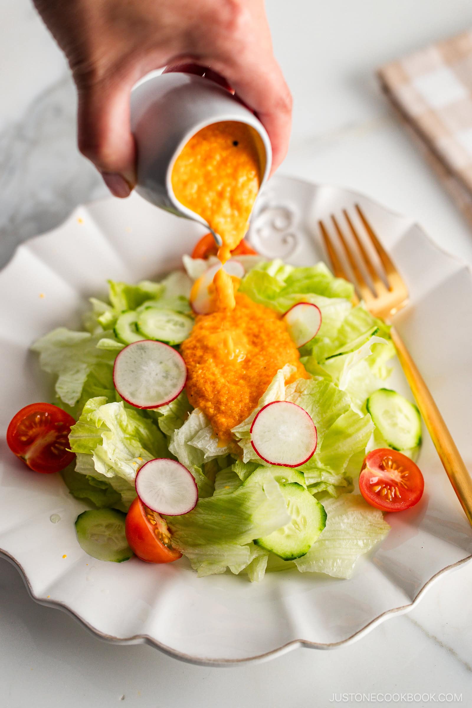 A white plate of fresh green salad with lettuce, cucumbers, radishes, and cherry tomatoes, topped with vibrant orange carrot ginger dressing and served with a gold fork on a marble surface.