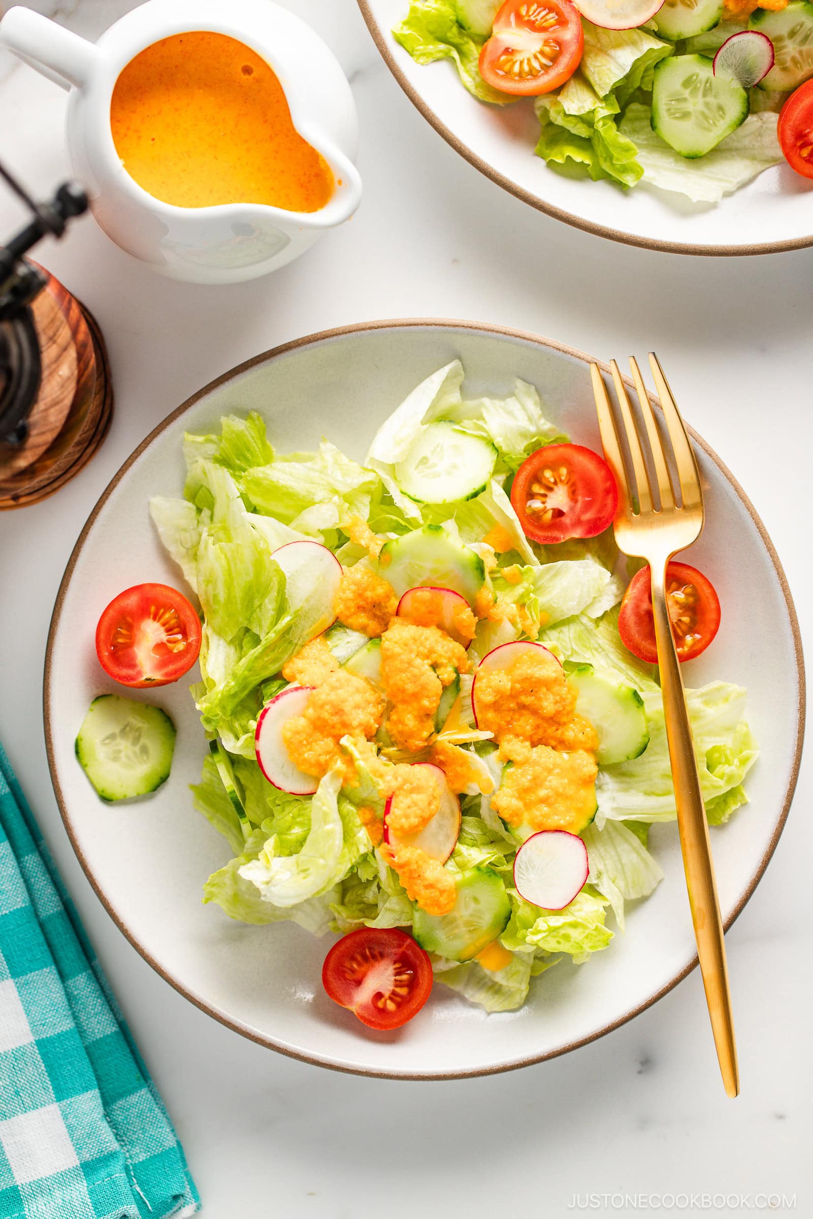 White plates of fresh green salad with lettuce, cucumbers, radishes, and cherry tomatoes, topped with vibrant orange carrot ginger dressing and served with a gold fork on a marble surface.