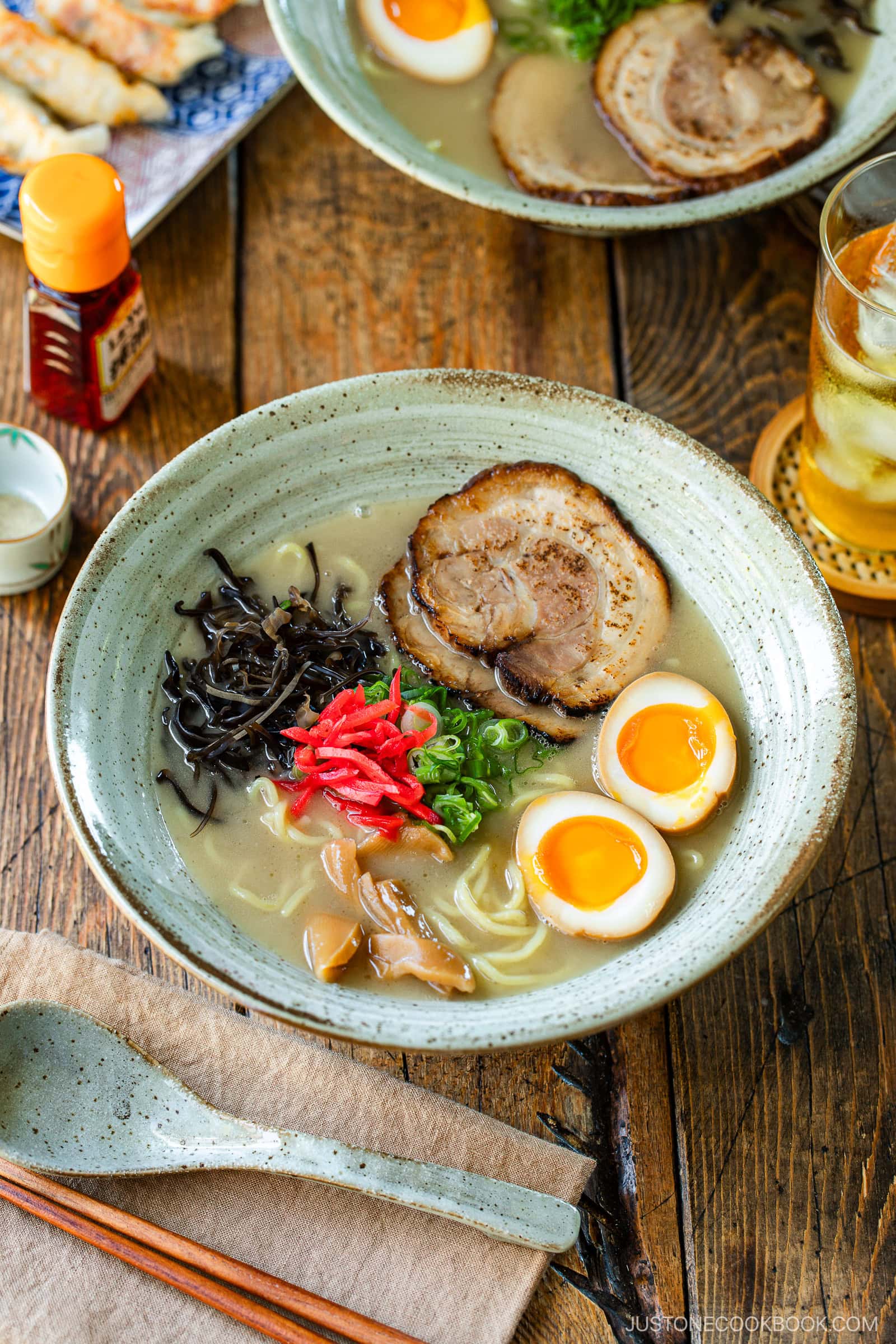 A rustic bowl containing noodles with tonkotsu broth, topped with chashu, ramen eggs, menma, kikurage, ginger, and chopped green onion. On the table, a plate of crispy gyoza and a glass of mugicha are served together.