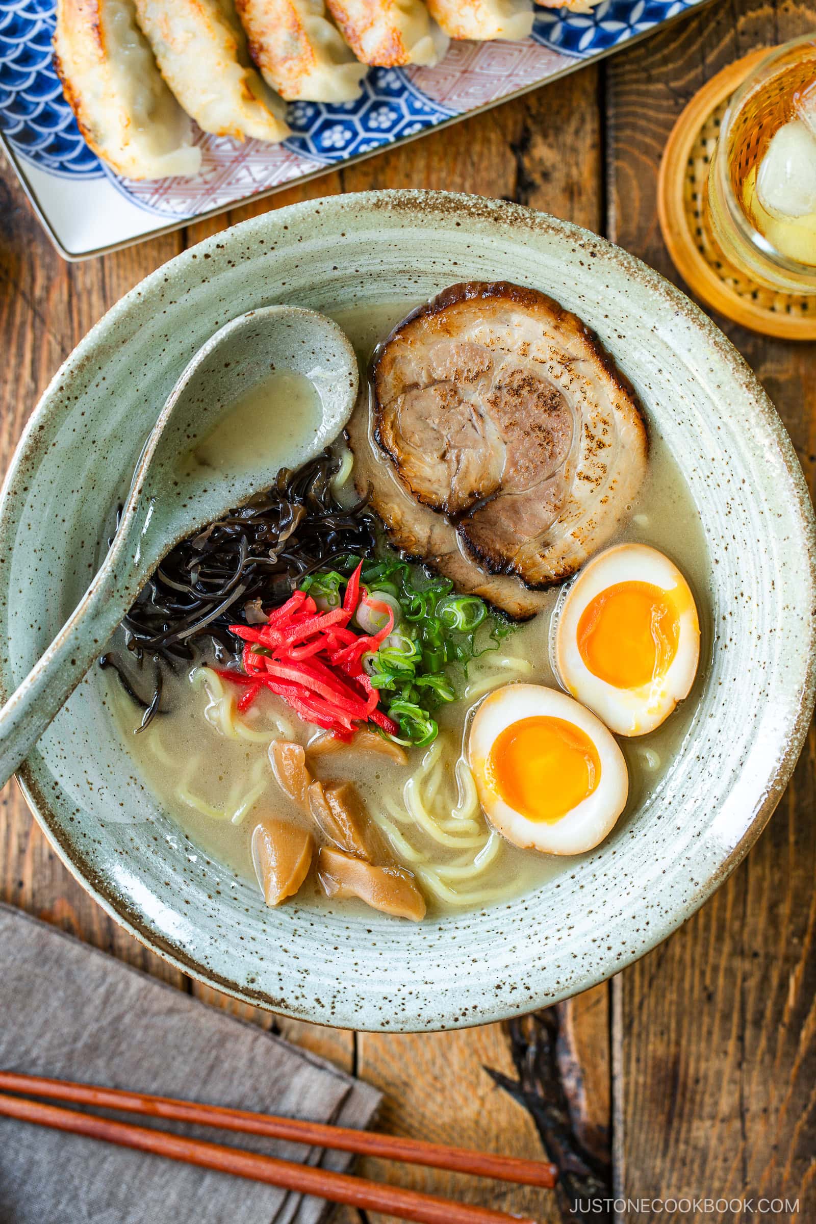 A rustic bowl containing noodles with tonkotsu broth, topped with chashu, ramen eggs, menma, kikurage, ginger, and chopped green onion. On the table, a plate of crispy gyoza and a glass of mugicha are served together.
